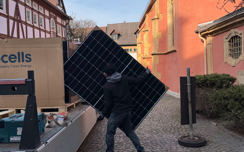 Ein Arbeiter trägt ein Photovoltaik-Modul auf einem gepflasterten Weg neben einem Lieferwagen.