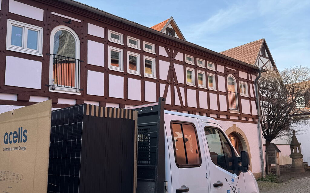 Ein Transporter steht vor einem Fachwerkhaus mit Photovoltaik-Panels auf der Ladefläche, bereit zur Installation.